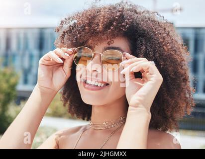 Portrait d'une jeune femme branchée de race mixte avec un afro souriant et posant seul à l'extérieur. Femme hispanique portant des lunettes de soleil et se sentant heureuse Banque D'Images