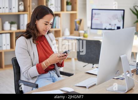 Jeune femme d'affaires asiatique envoyant des SMS sur un téléphone portable tout en travaillant sur un ordinateur de bureau à une table dans un bureau. Une femme qui vérifie uniquement les médias sociaux et Banque D'Images