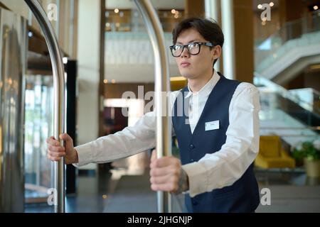 Jeune homme en uniforme de groom poussant chariot avec les bagages des clients de l'hôtel tout en se déplaçant le long du salon vers les chambres des clients Banque D'Images