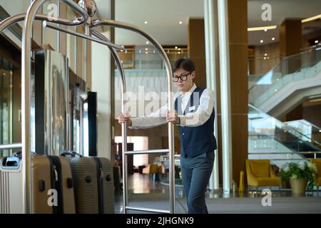 Un jeune groom en uniforme se déplaçant le long d'un couloir ou d'un salon traversant avec des valises de clients de l'hôtel ou de clients Banque D'Images