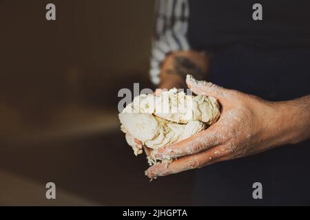 Un homme dans un tablier bleu tient la pâte de blé frais dans ses mains pour en faire une délicieuse pizza. Cuisine maison. Le processus de cuisson. Banque D'Images