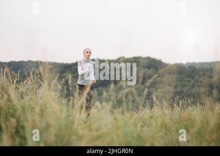 Bonne femme arabe dans hijab en course active au parc d'été. Jeune femme qui passe du temps libre sur l'air frais. Femme musulmane souriante qui fait du jogging pendant l'entraînement en plein air. Banque D'Images