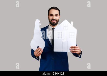 Homme barbu positif tenant maison de papier et grande clé, achat immobilier, services de location, hypothèque, portant le costume de style officiel. Prise de vue en studio isolée sur fond gris. Banque D'Images
