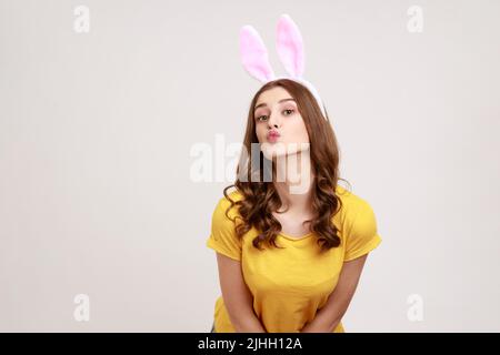 Drôle belle adolescente fille dans un style jaune décontracté T-shirt, debout et regardant l'appareil photo avec des oreilles de lapin, faisant le geste de bisou. Prise de vue en studio isolée sur fond gris. Banque D'Images