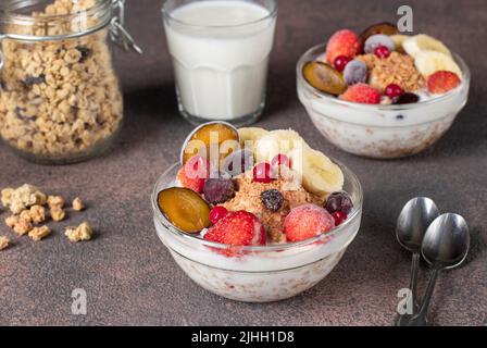 Muesli croustillants au granola avec yaourt naturel, baies glacées et fruits dans des bols en verre sur fond marron, petit déjeuner sain Banque D'Images