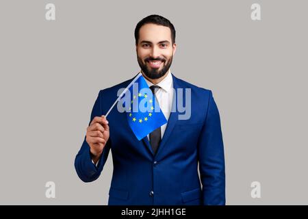 Citoyenneté dans l'Union européenne, immigration. Portrait d'un homme à barbe heureux montrant le drapeau de l'UE et souriant à l'appareil photo, portant un costume de style officiel. Prise de vue en studio isolée sur fond gris. Banque D'Images