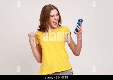 Portrait d'une adolescente surjoyée en T-shirt criant pour la joie, criant avec le téléphone mobile en main, clamant les poings, les gains réussis d'Internet. Prise de vue en studio isolée sur fond gris. Banque D'Images