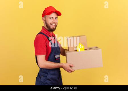 Portrait en profil de l'homme de livraison portant un uniforme bleu et un capuchon rouge tenant des boîtes en carton et une livraison porte-à-porte en toute sécurité dans les maisons en papier. Studio d'intérieur isolé sur fond jaune. Banque D'Images