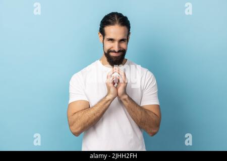 Portrait d'un homme impie avec une barbe portant un T-shirt blanc qui fume et conspirait en dérrant le plan, en pensant à une idée intelligente délicate, veut tricher. Studio d'intérieur isolé sur fond bleu. Banque D'Images