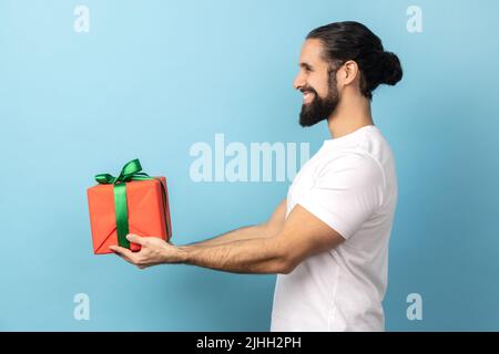Vue latérale d'un bel homme enchanté avec une barbe portant un T-shirt blanc donnant une boîte cadeau enveloppée et souriant, regardant à l'avance, des vacances heureuses. Studio d'intérieur isolé sur fond bleu. Banque D'Images