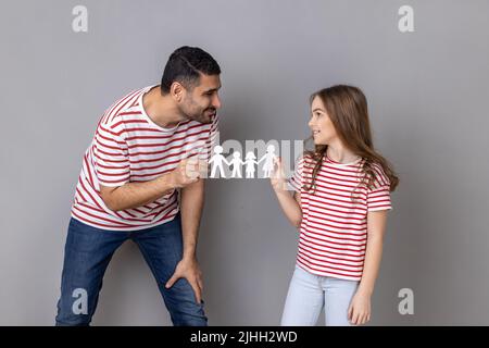 Portrait d'un père et d'une fille heureux et enchantés en T-shirts rayés, tenant la chaîne de personnes en papier, la relation familiale, l'enfance et la parentalité. Prise de vue en studio isolée sur fond gris. Banque D'Images