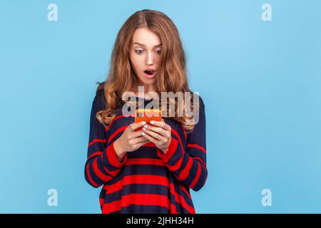 Portrait d'une femme surprise portant un chandail rayé de style décontracté, tenant le smartphone dans les mains, voit le contenu choquant, debout avec la bouche ouverte. Studio d'intérieur isolé sur fond bleu. Banque D'Images