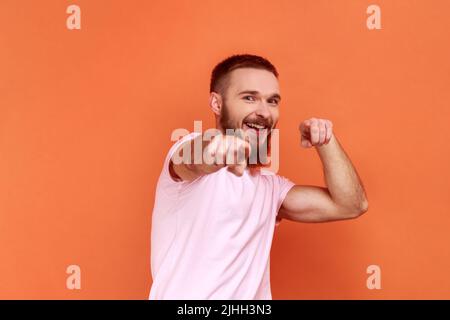 Portrait de beau barbu homme pointant vers l'appareil photo, souriant, faisant le choix heureux, nous avons besoin de vous concept, portant le T-shirt rose. Studio d'intérieur isolé sur fond orange. Banque D'Images