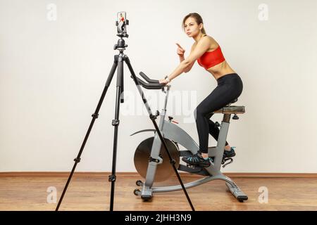 Portrait complet de femme sport blogger ayant entraînement cardio et cours en ligne en streaming, pointant le doigt vers l'appareil photo, portant des collants de sport et le haut rouge. Studio d'intérieur tourné sur fond de mur gris Banque D'Images