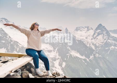 Bonne femme attrayante en lunettes de soleil se trouve sur un rocher avec des mains levées et regardant vers un ciel. Bon touriste dans les montagnes. Sommets enneigés à l'arrière Banque D'Images