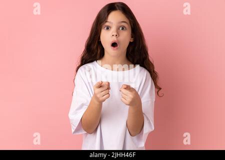 Portrait d'une petite fille stupéfaite portant un T-shirt blanc pointant vers l'appareil photo avec une expression surprise choquée, faisant le choix. Studio d'intérieur isolé sur fond rose. Banque D'Images