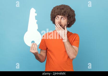 Portrait d'un homme choqué avec une coiffure afro portant un T-shirt orange tenant une clé en papier et couvrant la bouche, étant surpris, solution inattendue. Studio d'intérieur isolé sur fond bleu. Banque D'Images