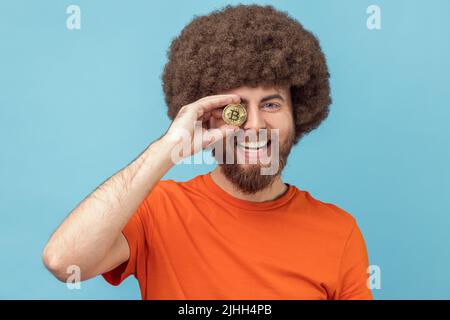 Portrait de l'homme optimiste avec la coiffure afro portant un T-shirt orange couvrant l'oeil avec le bitcoin d'or, ayant l'expression positive, la monnaie crypto. Studio d'intérieur isolé sur fond bleu. Banque D'Images