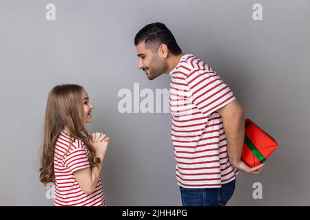 Portrait de père et fille heureux de fête en T-shirts à rayures, homme cachant boîte cadeau derrière son dos, petite fille attendant son cadeau. Prise de vue en studio isolée sur fond gris. Banque D'Images