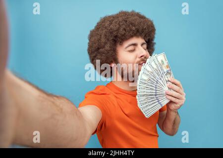 Portrait d'homme riche satisfait avec la coiffure afro portant un T-shirt orange tenant et sentant des billets de dollars, étant ravi d'obtenir salaire, POV. Studio d'intérieur isolé sur fond bleu. Banque D'Images