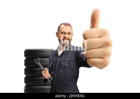 Mécanicien dans un uniforme tenant une clé et montrant les pouces vers le haut, les pneus de voiture dans le dos, isolé sur fond blanc Banque D'Images