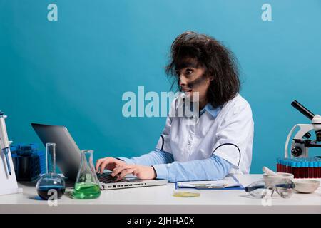 Maniaque goofy scientifique avec regard wacky en utilisant l'ordinateur pour analyser les composés chimiques. Un chimiste fou et fou avec un regard sauvage assis au bureau en laboratoire et analysant un rapport d'expérience raté sur un ordinateur portable Banque D'Images