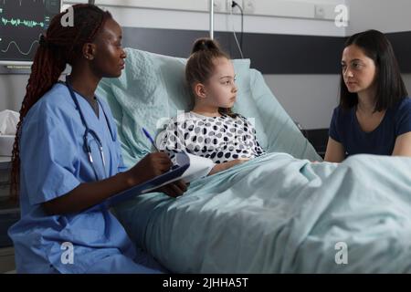 Une infirmière afro-américaine consultante une petite fille hospitalisée se reposant dans une clinique pédiatrique. Enfant malade se rétablissant de la maladie pendant que le personnel médical effectue un contrôle de santé. Banque D'Images