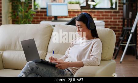 Femme asiatique concentrée prenant des notes tout en travaillant à distance et en écoutant de la musique à la maison dans le salon. Personne attentive travaillant à distance sur un ordinateur portable tout en rédigeant des informations importantes sur le poste. Banque D'Images