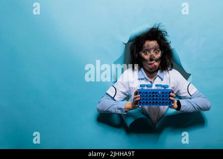 Un chimiste fou avec un aspect amusant et des cheveux enrasés qui collent la langue tout en ayant un rack rempli de tubes à essai en plastique. Scientifique fou au visage sale et à l'expression de goofy ayant stand rempli d'échantillons. Banque D'Images