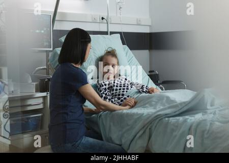 Mère assise à côté d'une fille malade hospitalisée se reposant dans un service pédiatrique de l'hôpital. Femme attentionnée réconfortant petite fille malade sous traitement dans la chambre de l'établissement de santé pour enfants. Banque D'Images