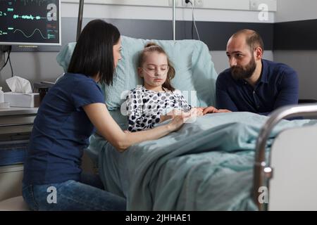 Parents inquiets assis à côté d'une fille malade hospitalisé dans un établissement de soins pédiatriques. Dormir une petite fille malade réconfortée par la mère et le père en s'occupant dans le lit du patient. Banque D'Images