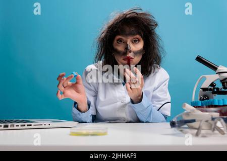 Un chimiste fou et fou qui sent le sérum rouge liquide expérimental tout en étant assis au bureau en laboratoire. Scientifique fou avec des cheveux wacky et sale face tenant tube rempli de composés chimiques. Banque D'Images