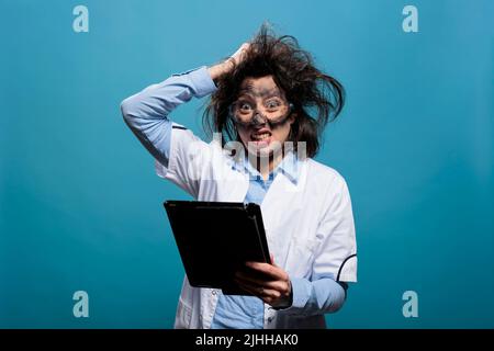 Un scientifique fou tire ses cheveux désordonnés après un échec de l'expérience chimique tout en ayant une tablette moderne. Travailleur de laboratoire lunatique avec écran tactile et visage sale enragé après l'explosion. Banque D'Images