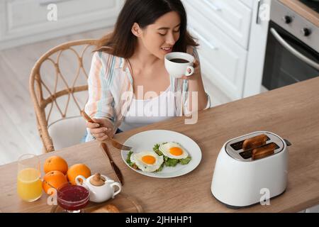 Belle jeune femme asiatique buvant du café et mangeant des toasts savoureux avec des œufs frits dans la cuisine Banque D'Images