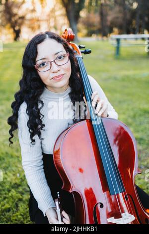 Bonne jeune femme brune avec des lunettes souriantes et jouant du violoncelle au coucher du soleil dans le parc. Verticale. Banque D'Images