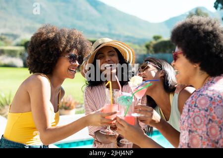 Des jeunes amies biraciales gaies toasting cocktails tout en s'amusant à la fête d'été au bord de la piscine Banque D'Images