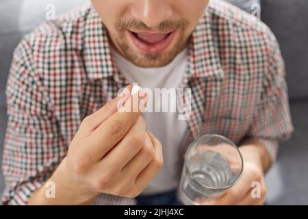 Malade jeune homme qui tient un verre d'eau dans sa main met une pilule dans sa bouche Banque D'Images