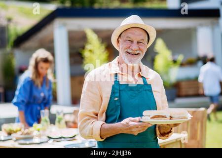 Joyeux homme senior servant des hamburgers lors de la fête de jardin de plusieurs générations en été. Banque D'Images
