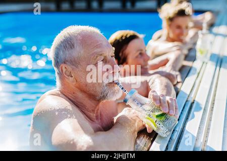 Homme senior qui apprécie la boisson froide en se tenant dans la piscine avec sa famille. Banque D'Images