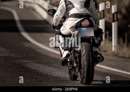 vue arrière d'une jeune motard vêtue d'un costume de protection en cuir et d'un casque de sécurité qui fait rouler sa moto sur une route asphaltée Banque D'Images