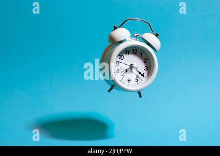 Réveil de style rétro blanc volant sur fond bleu jetant une ombre dure. Horloge analogique volante. Banque D'Images