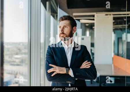 Homme d'affaires réussi en tenue formelle debout avec les bras pliés dans le bureau de collègue moderne et regardant par la fenêtre Banque D'Images