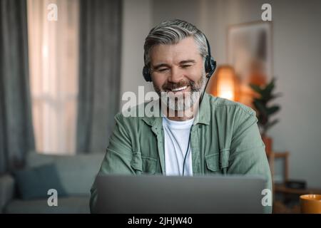 Un homme européen âgé souriant avec une barbe dans un casque regarde l'ordinateur portable, parle avec le client, regarde la leçon vidéo Banque D'Images