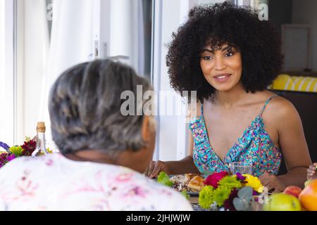 Jeune femme souriante multiraciale aux cheveux afro parlant avec une femme âgée à la table de salle à manger à la maison Banque D'Images