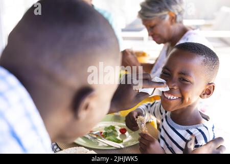 Un père multiracial nettoie le nez de son fils souriant avec du papier de soie tout en déjeunant à table Banque D'Images