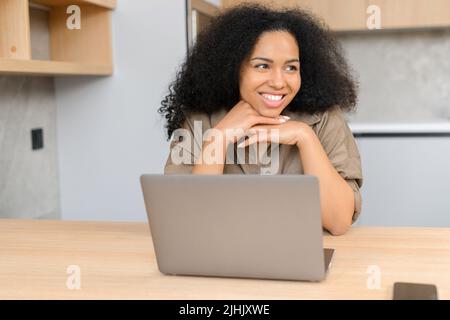 Portrait d'une jeune femme multiraciale assise au bureau avec un ordinateur portable, regardant loin, tout en envoyant un message à un ami, discutant avec un sourire agréable Banque D'Images