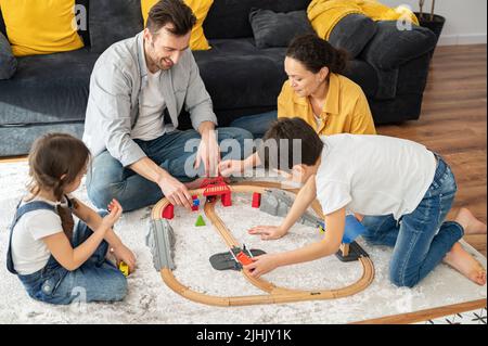 Une famille charmante de quatre personnes passe le week-end ensemble à la maison, parents et deux enfants jouant des jouets, le chemin de fer de construction de frères et sœurs, s'amuser. Maman, papa et enfants assis sur le sol dans un salon confortable et jouant Banque D'Images