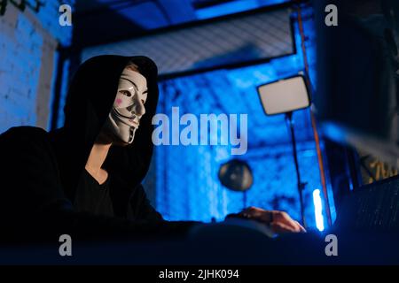 Gros plan de l'homme hacker à capuchon non reconnaissable portant un code de saisie de masque anonyme, cassant le mot de passe sur le clavier sans fil Banque D'Images