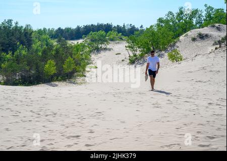 Un jeune homme de la vingtaine marchant pieds nus sur les dunes de sable transportant ses entraîneurs à Sandbanks Dunes Beach, comté de Prince Edward, Ontario, Canada. Banque D'Images