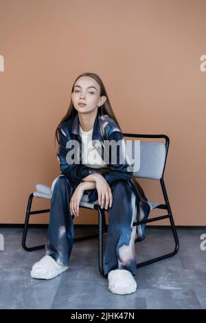 femme pleine longueur dans des vêtements tendance et des pantoufles moelleuses assis sur une chaise moderne sur beige Banque D'Images